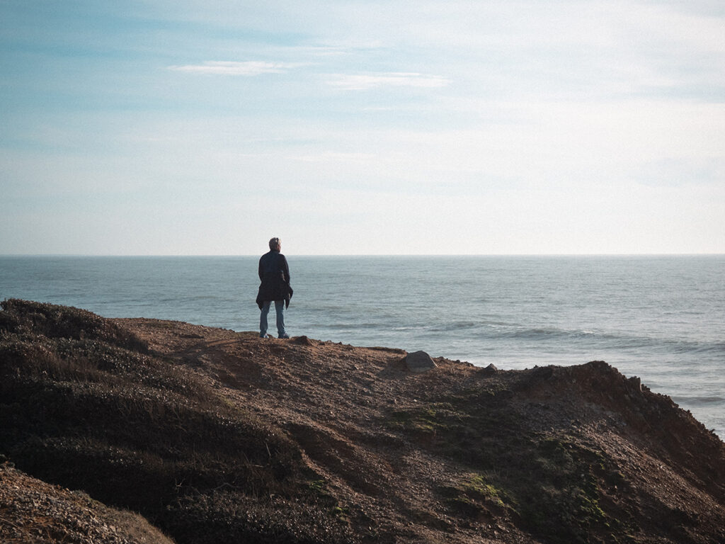 Homme debout face à l'océan, avec son manteau noué autour de la taille.