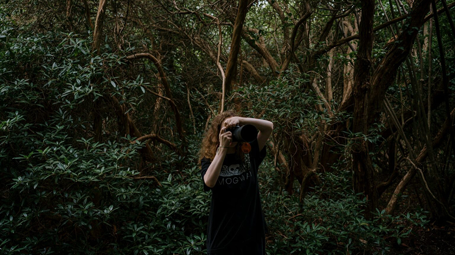 Une femme blanche tient son appareil photo prêt à déclencher. On ne voit pas son visage. Elle est dans la forêt.