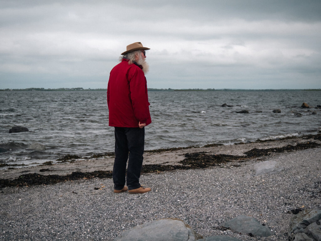Un homme âgé face à la mer. Il porte un coupe-vent rouge, un chapeau, des lunettes de soleil et une longue barbe blanche.