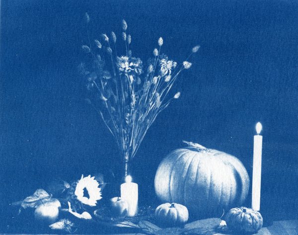 Photographie monochrome bleu qui représente une nature morte automnale. Sur une table, on trouve un bouquet de fleurs séchées, une grosse citrouille et deux petites, un tournesol, des pommes ainsi que deux bougies allumées.